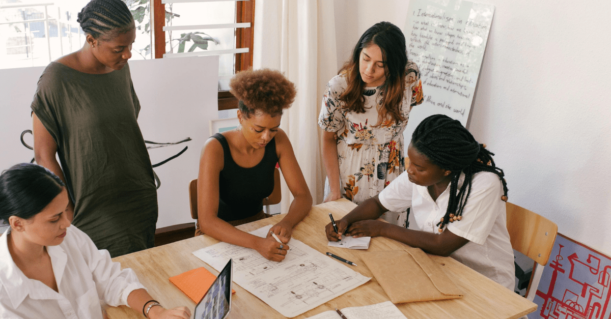 Cinco mulheres diversas sentadas em uma mesa escrevendo, conversando, mexendo no computador.